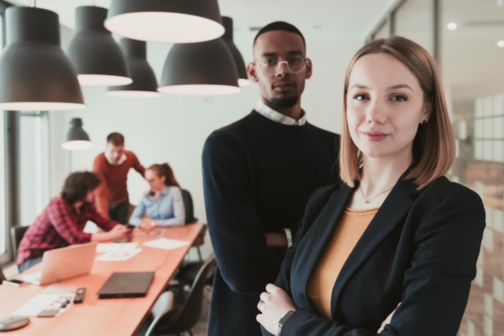 Confident young professionals standing in a modern office with coworkers collaborating in the background.